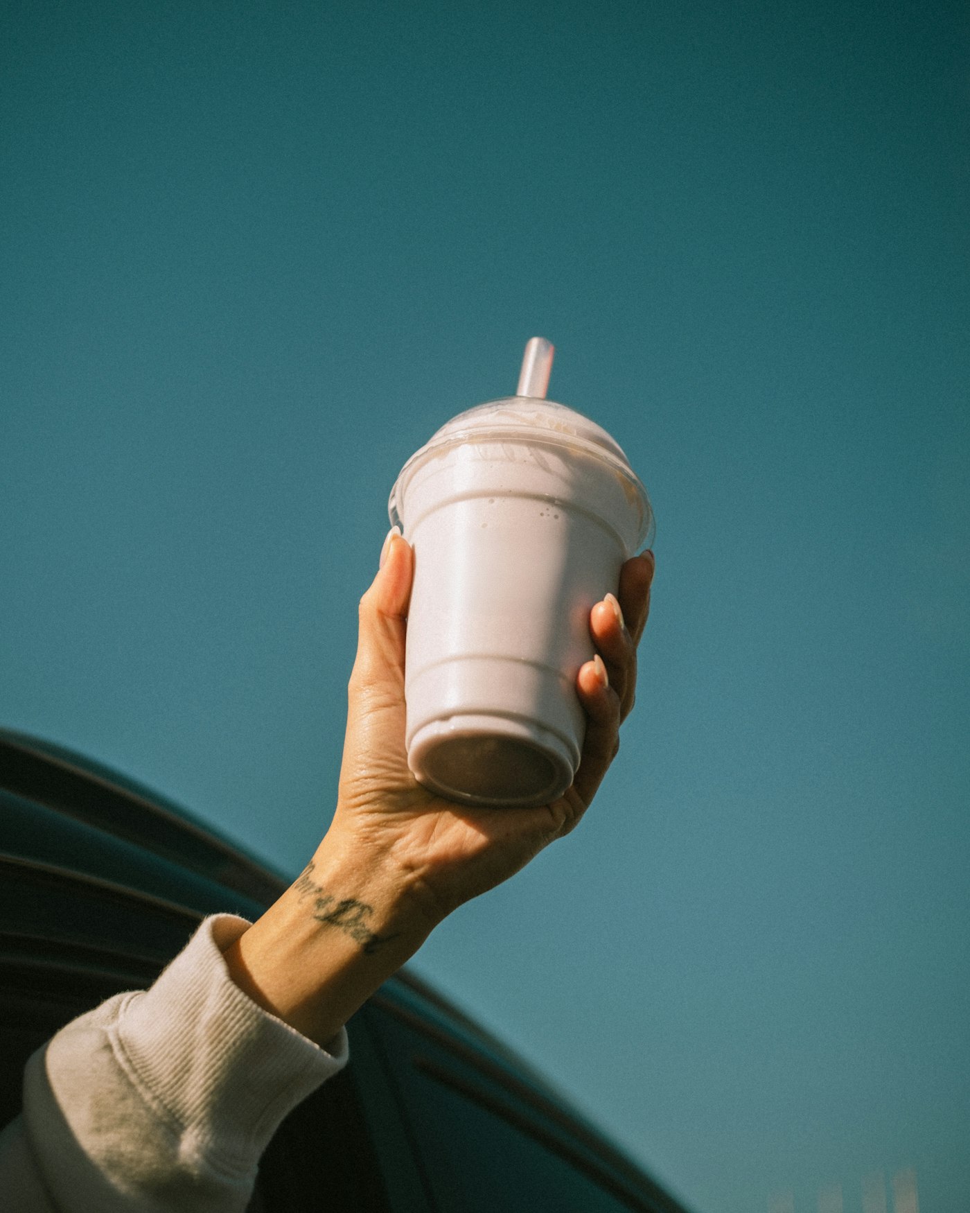A hand holding a takeaway shake out of a car window against a blue sky